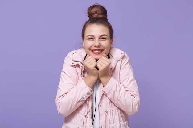 Eccitata ragazza affascinante con i capelli chignon, tenendo i pugni sotto il mento e sorridendo ampiamente, guardando direttamente la fotocamera, sognando qualcosa di fantastico, in posa isolato su sfondo lilla.