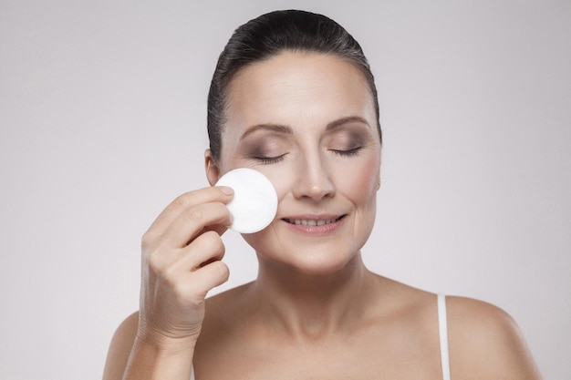 Excited cheerful confident lovely beautiful mature woman is using a cotton pad with micellar water for removing make up from face, isolated on grey background, copy space, studio shot, indoor