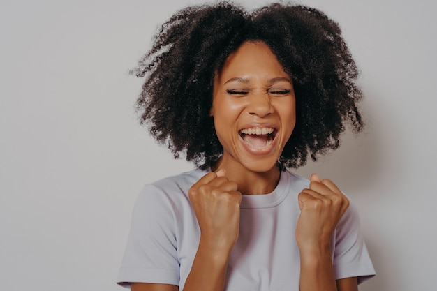 Eccitata ragazza dalla pelle scura allegra stringe i pugni mentre chiude gli occhi esclama con vittoria o desiderosa di vincere, donna eccitata vestita con abiti casual grida sì, isolata su bianco