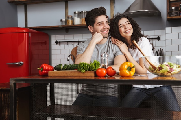 Взволнованная веселая молодая пара готовит полезный салат, сидя на кухне, глядя в сторону