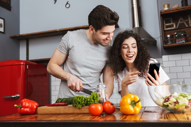Excited cheerful young couple cooking healthy salad while sitting at the kitchen, looking at mobile phone