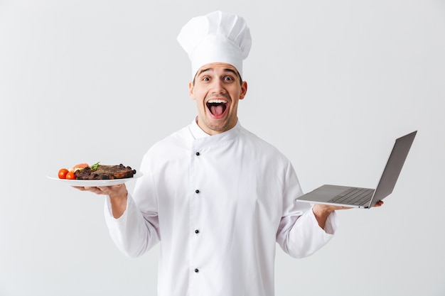 Excited chef cook wearing uniform standing over white wall, holding laptop computer, showing a dish