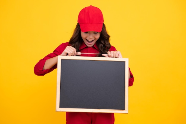 赤い制服とキャップで興奮した子供の女の子は黒板を保持します 10 代の少女は黄色の背景に分離された黒板を保持します