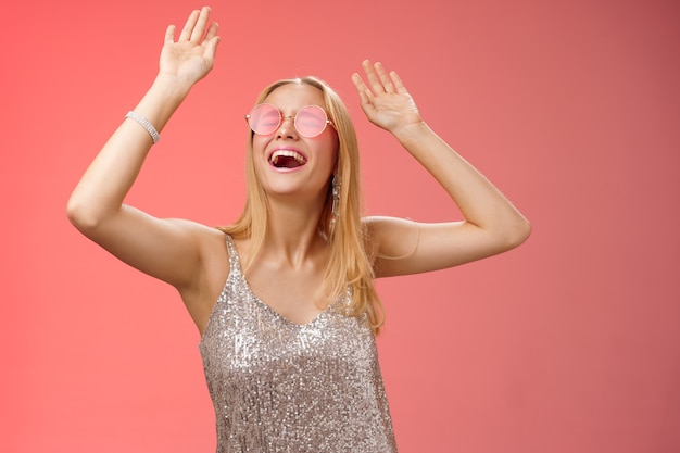 Excited chilling energized young blond woman in silver stylish glittering dress sunglasses raise hands up having fun dancing dance-floor nightclub throw party celebrate b-day, red background