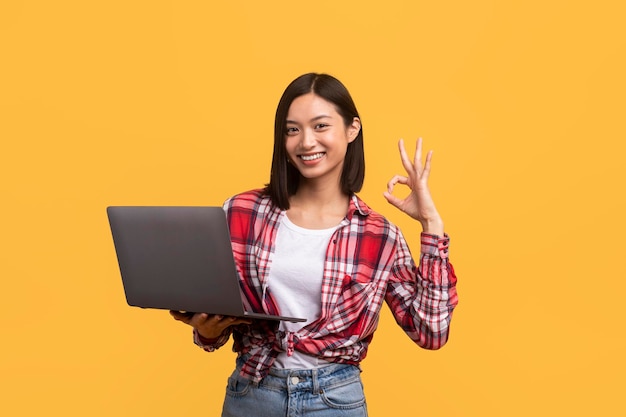 Excited chinese lady working on laptop and gesturing okay approving website standing over yellow background