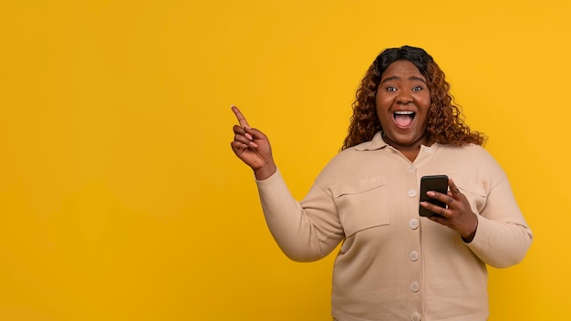 Photo excited chubby african american lady with smartphone pointing copy space