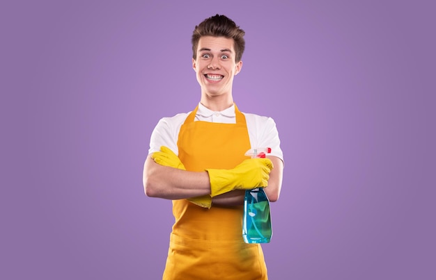 Excited comic male cleaner in apron and rubber gloves standing with liquid detergent on violet background in studio and looking at camera