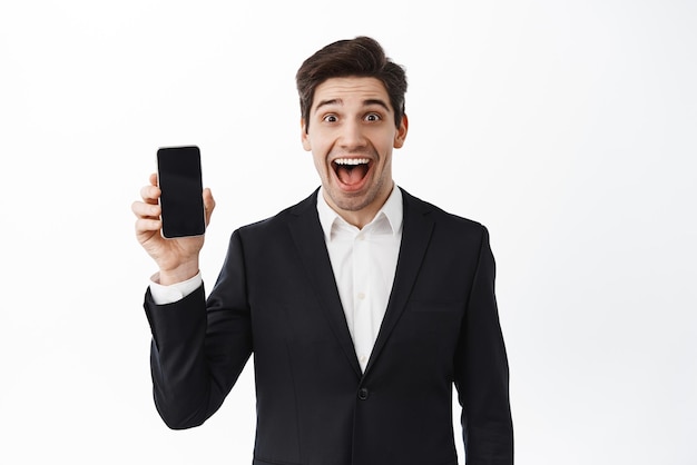 Excited corporate man introduce application showing empty phone screen and scream from excitement and joy standing in suit against white background