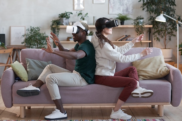 Photo excited couple of gamers in vr glasses sitting on sofa and playing virtual reality game in the room