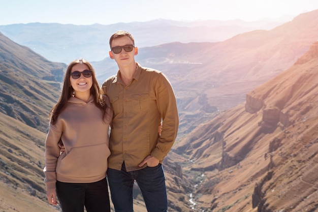 Excited couple in love hugs and smiles standing against large rocky brown mountains with canyon and small river under bright autumn sunlight