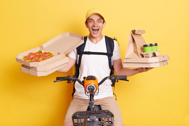 Foto corriere eccitato con zaino che indossa una maglietta bianca con scatole per pizza e caffè dal ristorante che urla con eccitazione in bicicletta isolata su sfondo giallo