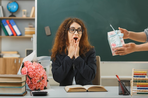 excited covered mouth with hands young female teacher received gift sitting at desk with school tools in classroom