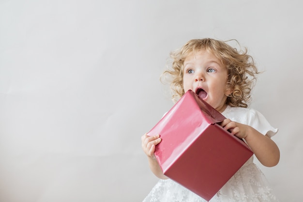 Excited curly girl in white dress with  gift on light gray wall