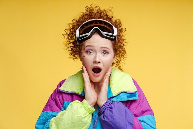 Excited curly woman wearing protective ski goggles ski overalls looking at camera isolated