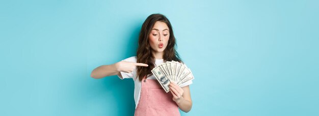 Excited cute girl holding money and pointing at dollar bills going on shopping standing over blue ba