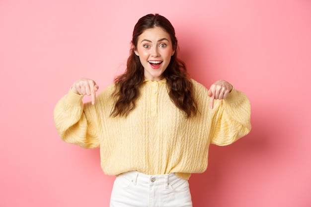 Excited cute girl showing advertisement pointing fingers down and smiling at camera demonstrating logo standing against pink wall