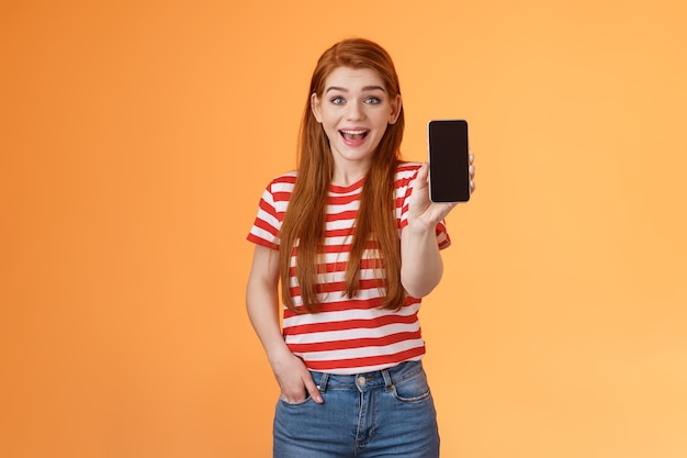 Excited cute outgoing redhead female impressed showing app hold smartphone introduce gadget feature ...