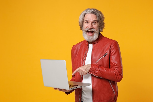 Excited elderly gray-haired mustache bearded man in red leather jacket posing isolated on yellow background. People lifestyle concept. Mock up copy space. Pointing index finger on laptop pc computer.