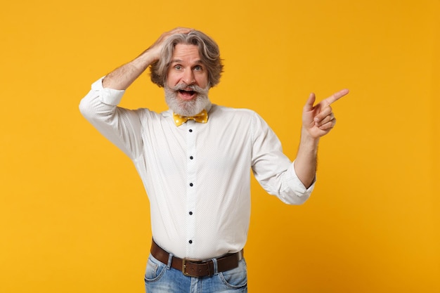 Excited elderly gray-haired mustache bearded man in white shirt\
bow tie posing isolated on yellow background. people lifestyle\
concept. mock up copy space. pointing finger aside, putting hand on\
head.