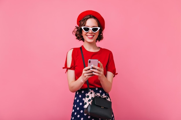 Excited european woman in elegant french outfit texting message Adorable caucasian girl in sunglasses and beret posing with phone