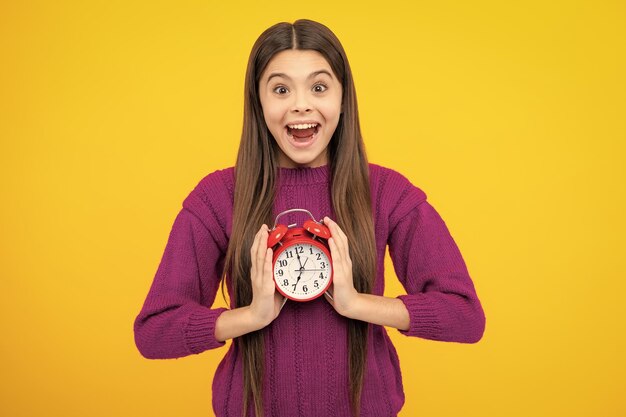 興奮した顔 10 代の学生の女の子は、黄色の背景に分離された時計を保持します 学校への時間