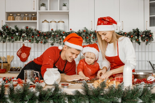 サンタの帽子をかぶった興奮した家族の母、父、息子がクリスマスライトの上に散らかったキッチンテーブルでクッキーカッターで生地を切る