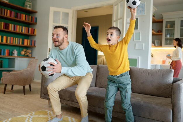 興奮した父と幼い息子が、自宅の居間のソファでサッカーの試合を応援し、見ている。素晴らしい時間を一緒に過ごす幸せな家族