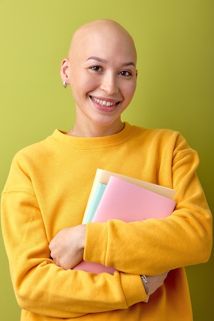 Excited female with bald head holding books in hands and\
smiling enjoying education studying hairless lady in casual yellow\
shirt posing at camera university concept people lifestyle