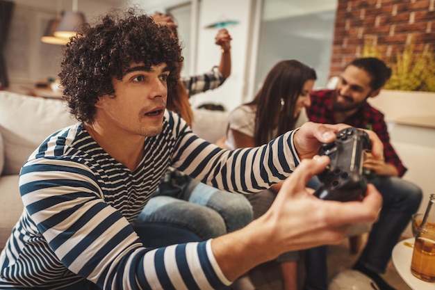 Excited friends are playing video games at home sitting on sofa with beer. They have great weekend in nice company indoors.