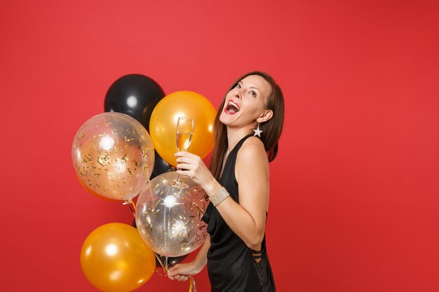 Excited funny girl in black dress celebrate looking up hold glass of champagne, air balloons isolated on red background. International Women's Day Happy New Year birthday mockup holiday party concept.