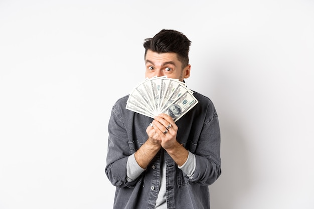 Возбужденный смешной парень прячет лицо за долларовыми купюрами и улыбается, показывая деньги наличными, стоя на белом фоне.