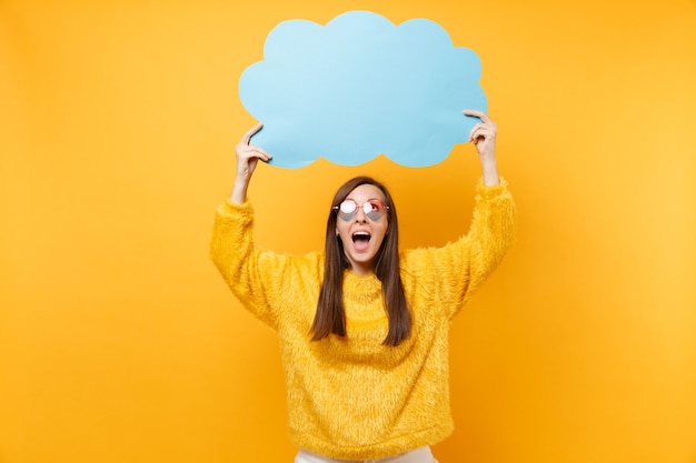 Excited funny young woman in heart eyeglasses holding empty blank blue say cloud, speech bubble isolated on bright yellow background. people sincere emotions, lifestyle concept. advertising area