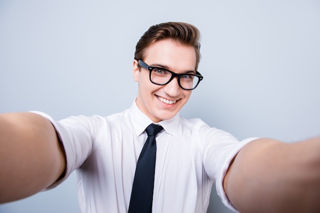 Excited geek young man in trendy glasses and formal wear is making selfie shot on camera, standing on a pure space, showing thumb up sign, smiling