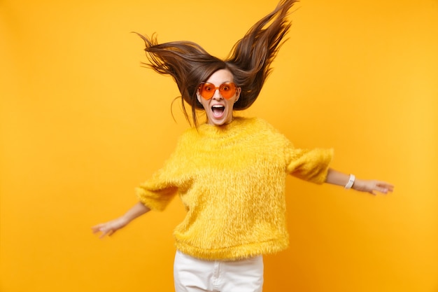 Excited girl in fur sweater and heart orange glasses fooling around in studio jump with flying hair isolated on bright yellow background. People sincere emotions, lifestyle concept. Advertising area.