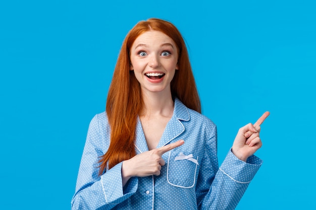 Excited girl hearing wonderful news, special discounts for holiday season. Attractive redhead female teenager nightwear smiling astonished and delighted, pointing upper right corner, blue wall