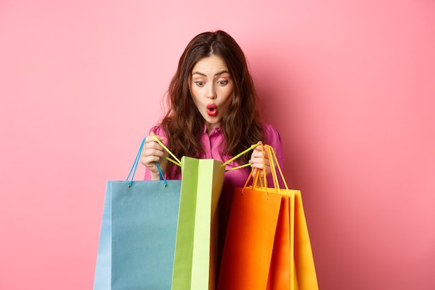 흥분된 소녀는 쇼핑 가방을 열고 깜짝 놀라며 행복한 얼굴로 선물을 확인하고 분홍색 벽에 서 있습니다. 공간 복사