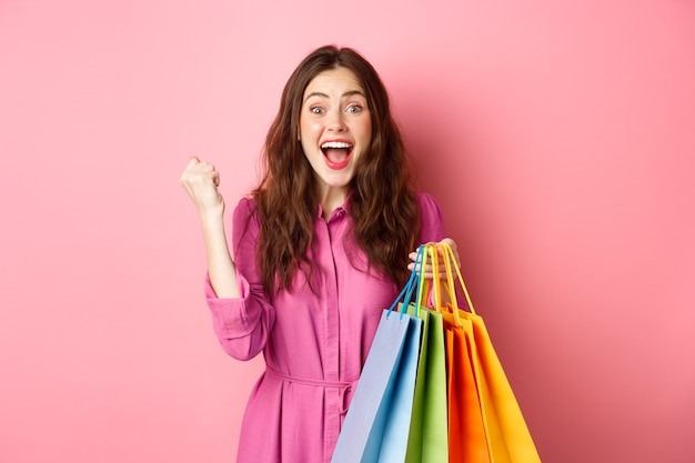 Excited girl scream of joy, making fist pump, holding shopping bag and rejoicing, standing in dress over pink wall.