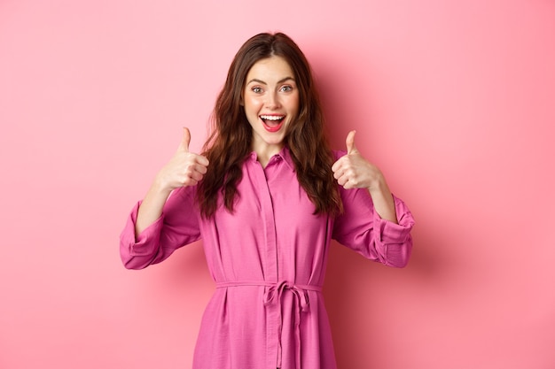 Excited girl showing thumbs up and smiling amazed, praise awesome product, standing against pink wall.