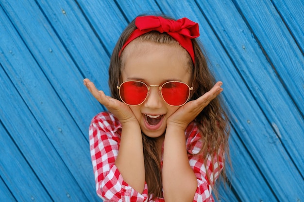 Photo excited girl in sunglasses opened mouth in surprise, putting hands to her cheeks