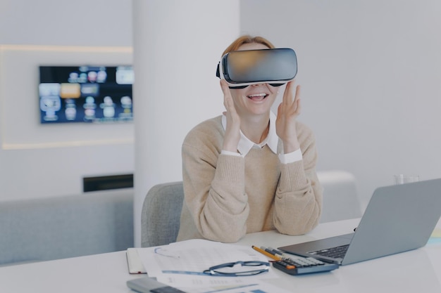 Excited girl in VR headset sitting at the desk in front of laptop Working on project in cyberspace