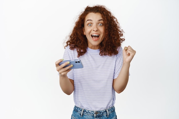 Excited girl winner, redhead woman holding mobile phone and looking amazed as if winning, become champion in video game, white background. Copy space