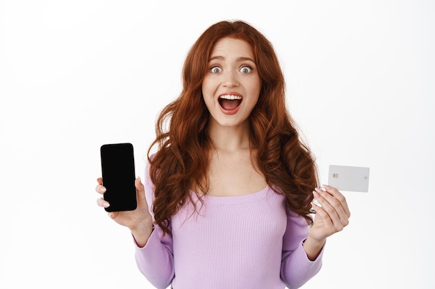 Excited girl with red curly hair, gasping wondered and amazed, showing smartphone blank screen and credit card, demonstrating shop online or bank promotion, standing against white background