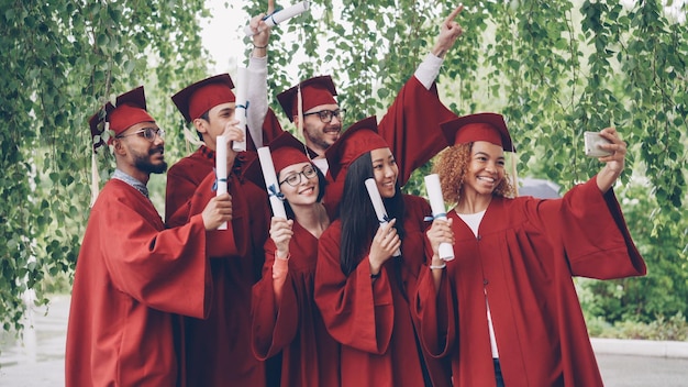 Excited graduating students are taking selfie with smartphone\
young people are waving diplomas posing smiling and laughing\
education and success concept