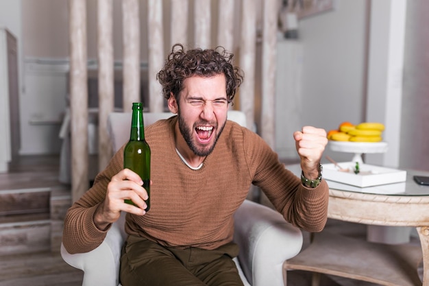 소파에 앉아 맥주를 마시고 축구를 보는 흥분된 남자. 스포츠, 행복, 그리고 사람들의 개념 - 웃는 남자가 TV로 스포츠를 보고 집에서 팀을 지원합니다. 집에서 축구를 보는 남자