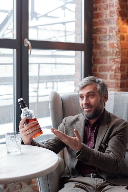 Excited handsome gray-haired man sitting in luxurious armchair and recommending whisky to friend