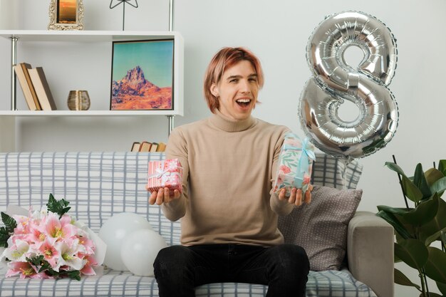 Excited handsome guy on happy women day holding present. sitting on sofa in living room