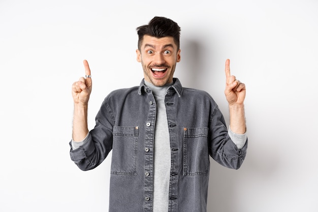 Excited handsome guy pointing fingers up, smiling happy and showing special deal, standing on white background.