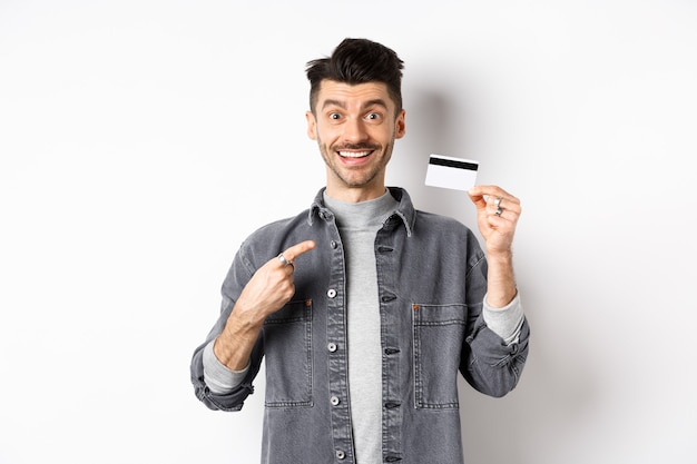 Excited handsome guy with moustache pointing finger at plastic credit card, smiling pleased, recommend good deal, standing on white background.