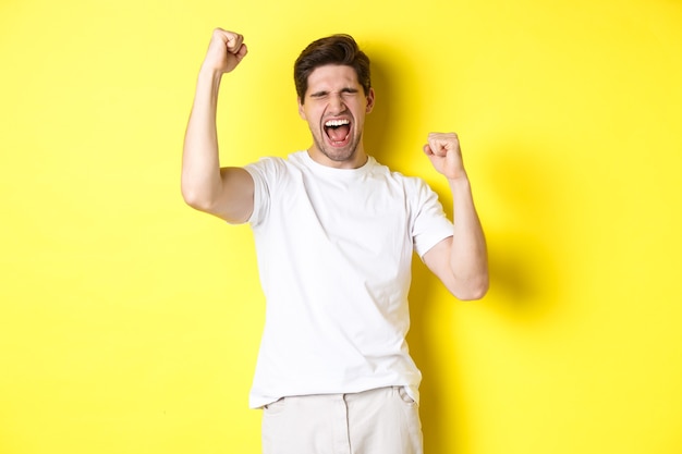 Excited handsome man winning and shouting for joy, raising hand up and triumping, achieve goal, standing over yellow wall