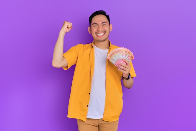 Excited handsome young Asian man holding money banknotes and celebrating success isolated on purple background